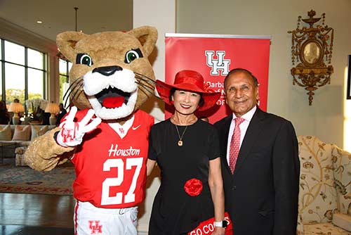A cougar mascot in a red and white football uniform stands next to an Indian woman and man wearing black formal wear.