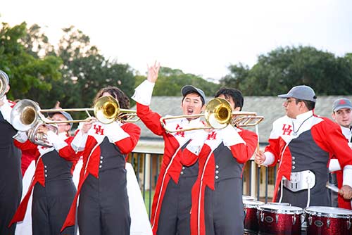 A college marching band in black, red and white uniforms play horn instruments and drums together.