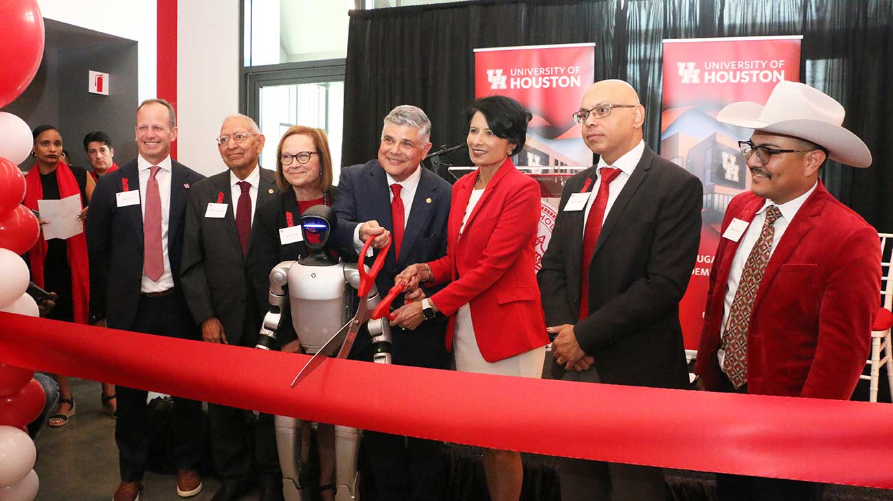 A group of men and women in business attire stand in front of large red ribbon. A man and woman next to a standing robot in the middle hold a pair of large scissors to cut the ribbon.