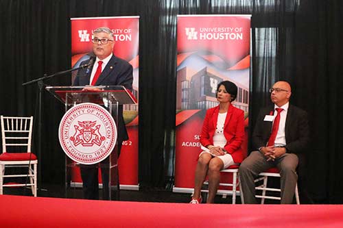 A man in a business suit stands at a podium giving a speech while a woman and another man sit in chairs next to him.