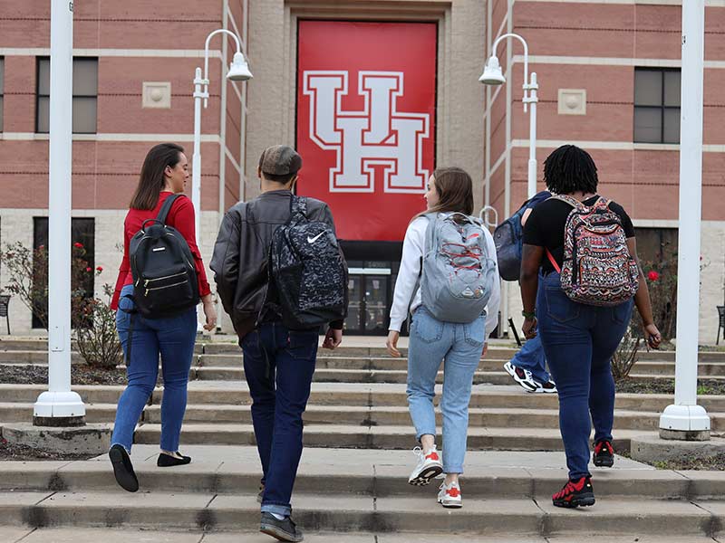 Four students walk towards the front entrance of a brick building with a large red UH banner.