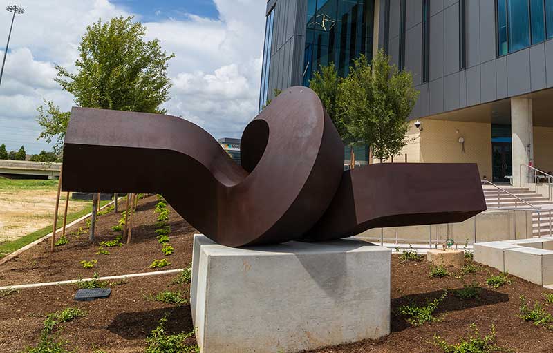 Photograph of a metal sculpture. Side view of a rectangular prism sits parallel to the ground and then twists in a loop on pedestal.