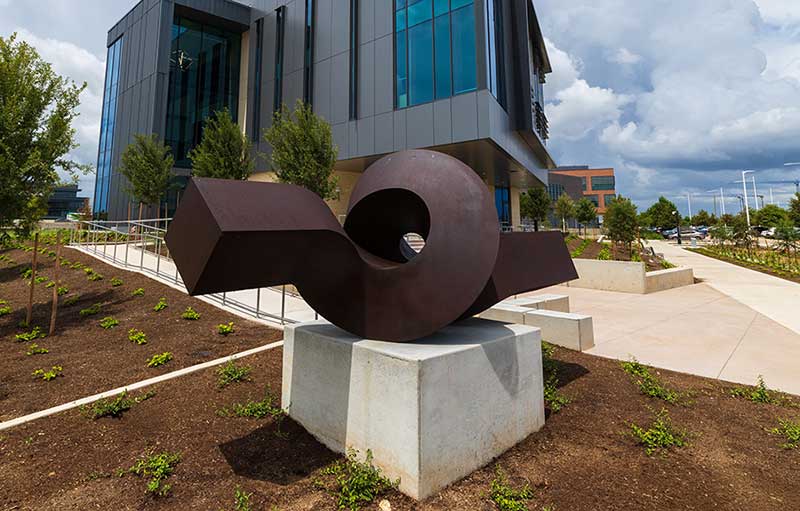 Photograph of a metal sculpture. A rectangular prism sits parallel to the ground and then twists in a loop on pedestal in front of a building.