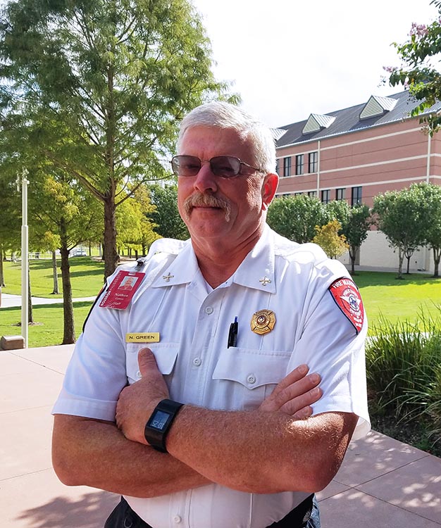 A man with grey hair and mustache wearing a white fire marshal uniform stands in front of building with trees.