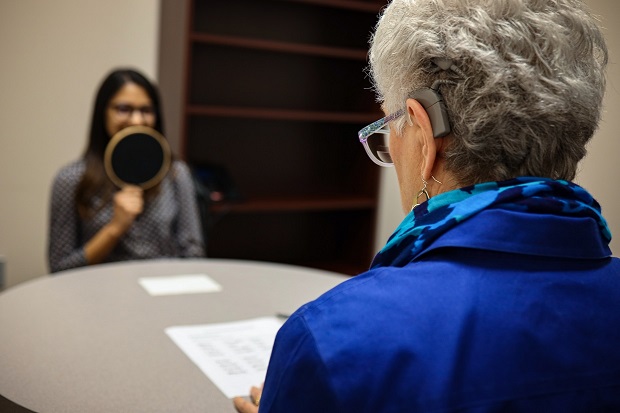 Portrait of the back of a silver-haired woman in a blue coat. A dark-haird woman with glasses covering mouth with a large round object.