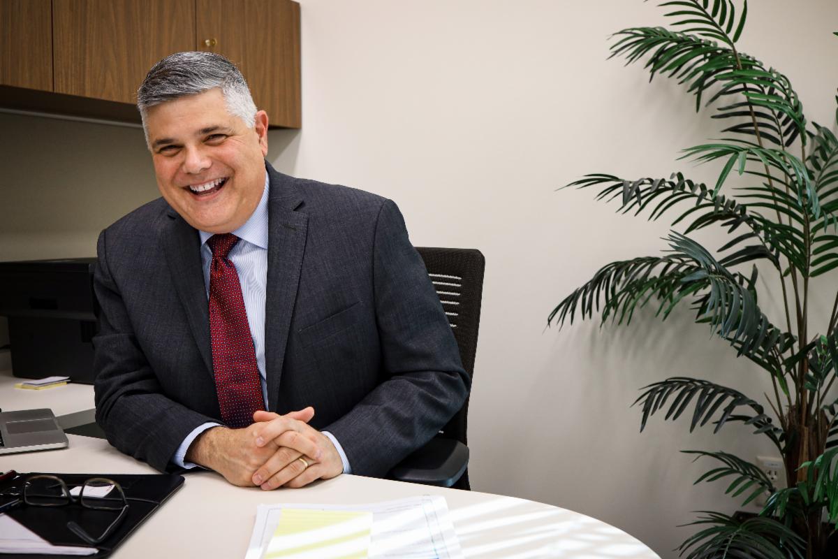 Portrait of a man laughing with short gray hair wearing a dark suit with a marron tie.