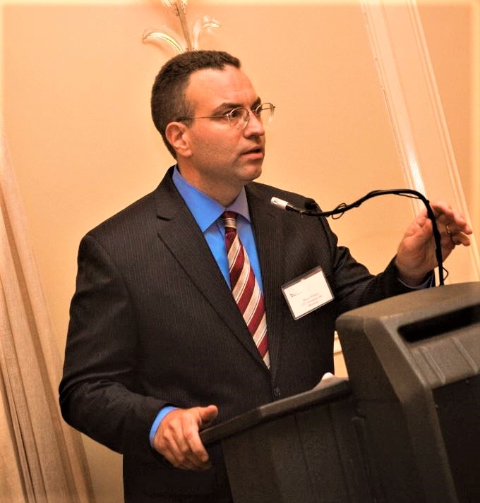 Portrait of a man in glasses wearing a black suit speaking into a microphone at a podium