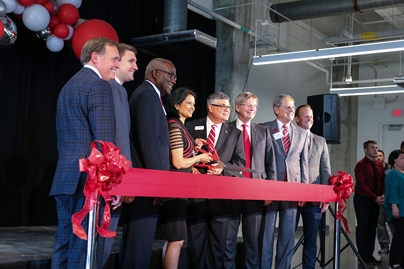 A group of men in suits stand around a man and woman holding a large open pair of scissors over a large red ribbon