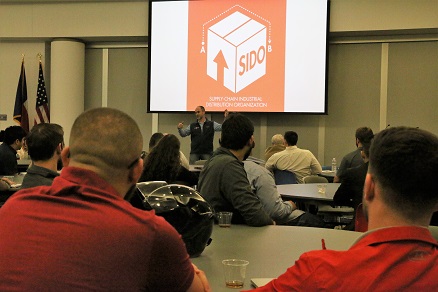 Students sitting at tables watching a man present in front of a projection screen