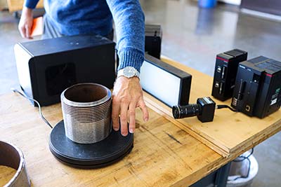 A person reaches for a silver cylindrical piece of electronic equipment surrounded by black pieces of electronic equipment sitting on a table.