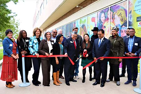 A group of women and men stand behind a red ribbon as a women with large scissors cuts the ribbon.