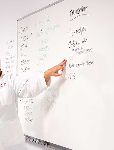 A faculty member in a white lab coat points to writing on a white board.