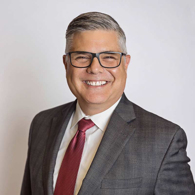 A man wearing a grey suit with a red tied and horn-rimmed glasses.