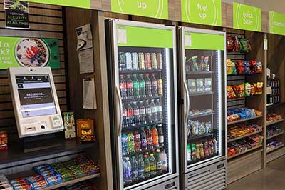 Shelves in a convenience store stocked with food and drinks next to a self-checkout kiosk..