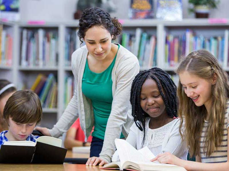 A teacher overlooks students reading books together at a desk in a library.
