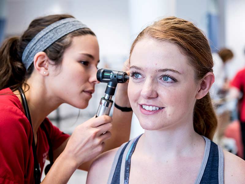 A nursing student holds an otoscope to a patient's ear in a hospital-style nursing lab.