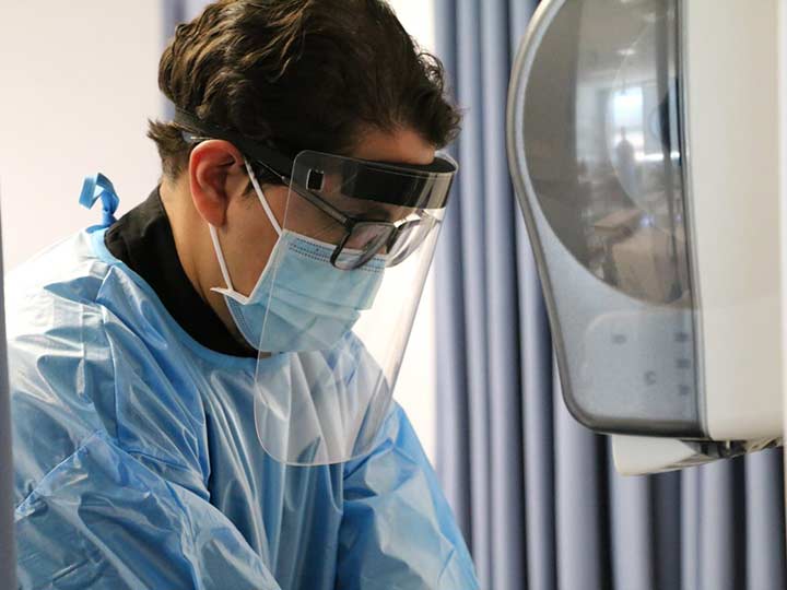 A nursing student wearing a face shield and gown while looking downward at a sink