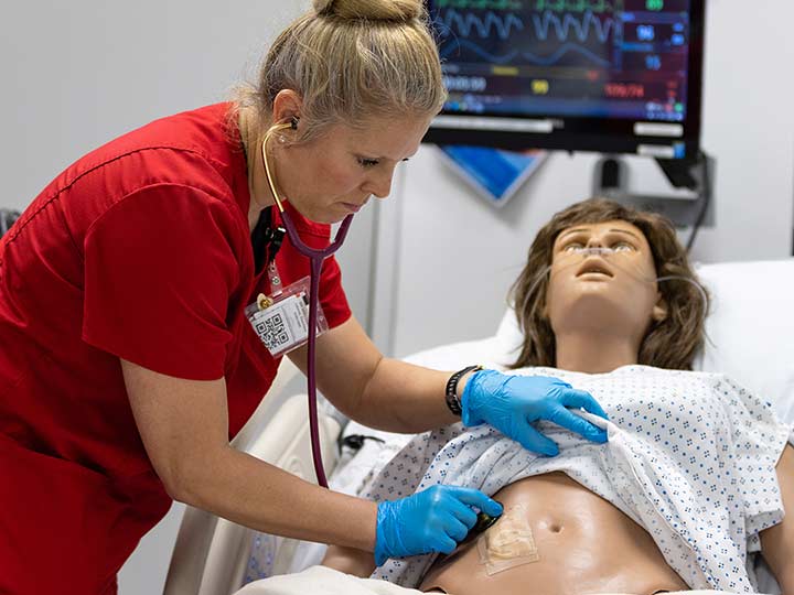 A nursing student places a stethoscope on the abdomen of a high-fidelity manikin with a bandaged wound.